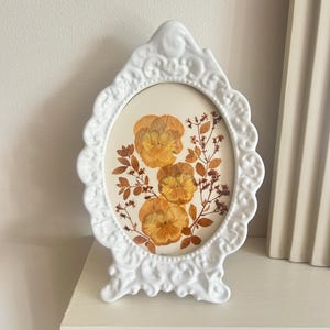 May include: White, ornate oval picture frame with pressed yellow and orange flowers. The frame has a decorative, scrolled design and stands on a white surface. The flowers are arranged with brown stems and leaves.