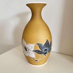 Peut inclure: Un petit vase en céramique brun avec un bord blanc et un motif floral. Le vase présente deux fleurs stylisées, une blanche et une noire, avec des contours blancs.