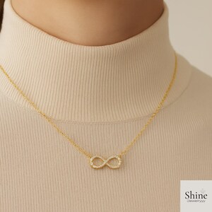 May include: A gold necklace with an infinity symbol pendant, set with small, clear gemstones. The necklace is shown against a cream-coloured turtleneck. The brand name "Shine Jewelryyy" is visible in the bottom right corner.