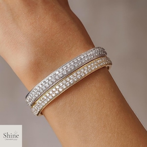 Peut inclure: Deux bracelets rigides, un argenté et un doré, ornés de plusieurs rangées de cristaux étincelants. Les bracelets sont présentés sur un poignet, mettant en valeur le design et l'éclat des bijoux. La marque "Shine Jewelryyy" est visible.