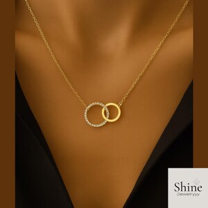 May include: A gold-colored necklace featuring two interlocking circles. One circle is encrusted with small, clear gemstones, while the other is a plain gold band. The necklace hangs on a delicate chain.