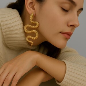 May include: Gold-colored snake earrings. The earrings are in the shape of a snake with textured scales. The snake's head is at the top, and the body curves down. The earrings are worn by a person wearing a cream-colored sweater.