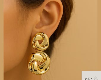 May include: Gold-colored, double-drop earrings with a knotted design. The earrings are shiny and reflect light, creating a bold, eye-catching accessory. The earrings are a statement piece of jewelry.
