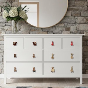 Op de afbeelding: Een witte dressoir met negen lades, elk versierd met knoppen in de vorm van dieren. De knoppen omvatten een luiaard, een beer, een giraffe en een konijn. Een vaas met witte bloemen en een ronde spiegel staan op de achtergrond.