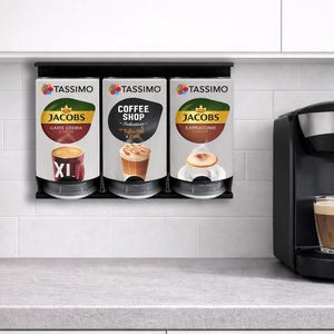 Op de afbeelding: Een zwarte Tassimo koffiemachine met een glazen mok gevuld met koffie. Drie Tassimo koffiepads zijn aan de muur bevestigd. De pads zijn gelabeld met Jacobs Caffe Crema, Coffee Shop en Jacobs Cappuccino. Een kleine plant in een pot staat op het aanrecht.