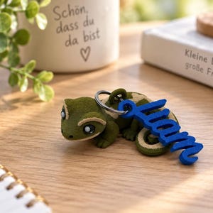 Könnte beinhalten: Ein Schlüsselanhänger in Form eines grünen und beigen Geckos mit einem Metallring, an dem ein leuchtend blaues Namensschild in Schreibschrift mit der Aufschrift Liam befestigt ist. Der Gecko hat große, dunkle Augen und eine strukturierte Oberfläche.