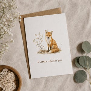 Può includere: Un biglietto d'auguri bianco con un'illustrazione ad acquerello di una volpe arancione e bianca seduta accanto a una piccola pianta. Il testo "a little note for you" è stampato in inchiostro marrone sotto la volpe. Il biglietto è in una busta marrone.