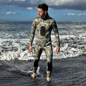 Könnte beinhalten: Eine Person trägt einen Tarnanzug mit einer schwarzen Kapuze und dem Wort "ANTARES" auf der Brust. Die Person steht im Wasser an einem Strand.