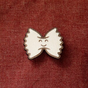 Peut inclure: Une broche en bois clair en forme de nœud papillon de pâtes farfalle avec un bord brun foncé. La surface de la pièce en bois présente un dessin au trait simple et gravé d'un visage souriant avec des yeux fermés et une petite bouche courbée.