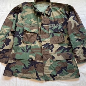 May include: A long-sleeve camouflage jacket featuring a woodland pattern of green, brown, and black. The garment includes four front pockets with flaps, a collar, and the text U.S. ARMY embroidered above the right chest pocket with shoulder patches.