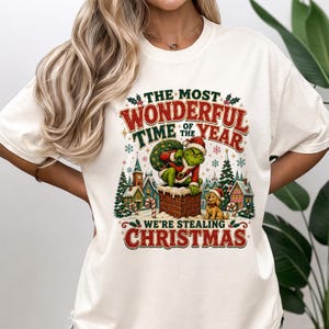 May include: A cream-colored t-shirt featuring a graphic print of a green character in a Santa suit emerging from a brick chimney with a sack. The text reads, "THE MOST WONDERFUL TIME OF THE YEAR WE'RE STEALING CHRISTMAS" amidst snowy houses and trees.
