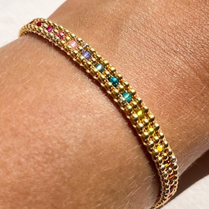 May include: Gold beaded bracelet with a rainbow of colorful beads. The bracelet is made of small, round gold beads and features a row of colorful beads in red, pink, yellow, green, and blue. The bracelet is worn on a wrist.