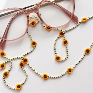 May include: Pink-framed glasses with a beaded chain featuring sunflower beads. The chain has yellow, brown, white, gold, and green beads. The glasses and chain are displayed on a white background. The chain is approximately 60 cm long.