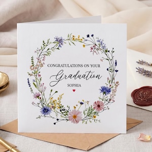 May include: A white graduation card with a floral wreath design. The card reads "Congratulations on your Graduation Sophia" in elegant script. A gold ring sits in a small gold dish.