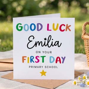 Puede incluir: Una tarjeta de felicitación cuadrada blanca con texto colorido que dice GOOD LUCK en letras de arcoíris, seguido del nombre Emilia en letra cursiva negra con una pequeña estrella, y el texto ON YOUR FIRST DAY PRIMARY SCHOOL en negro con una estrella amarilla.