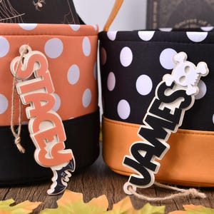May include: Two Halloween storage bins, one orange with white polka dots, the other black with white polka dots. Each has a name tag. The orange bin has a tag that says "STACEY" and the black bin says "JAMES".