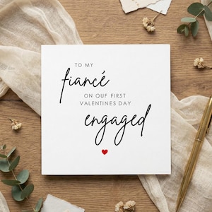 May include: A white card with the words "To My Fiance" and "Engaged" in elegant black script, with a small red heart. The card is on a wooden surface, accompanied by a gold pen and decorative elements.