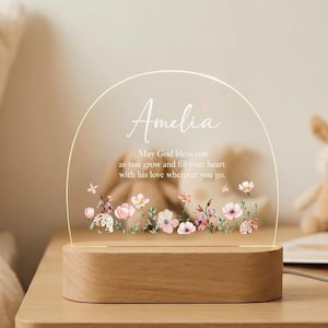 Puede incluir: Luz nocturna LED con base de madera y arco de acrílico transparente. El arco presenta el nombre "Amelia" y un diseño floral con flores rosas y blancas. El texto dice "May God bless you as you grow and fill your heart with his love wherever you go."