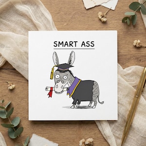 May include: A white square card featuring the text "SMART ASS" above a cartoon donkey in a graduation cap and gown, holding a scroll. The donkey is grey with black and purple details. The card is on a wooden surface.
