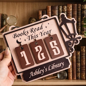 Puede incluir: Letrero de madera con el texto "Books Read This Year" y el número "125". El letrero tiene una linterna decorativa y está personalizado con "Ashley's Library". El fondo muestra una estantería con libros.