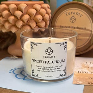 Op de afbeelding: Een glazen kandelaar met een crèmekleurige kaars. Het etiket luidt "Teramy Spiced Patchouli". De kaars is gemaakt met koolzaadwas en biologische essentiële oliën. Een houten lont is zichtbaar.