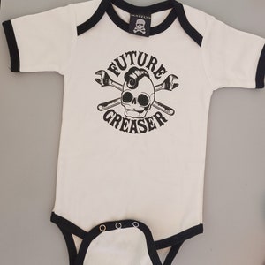 May include: White baby onesie with black trim and a black and white graphic of a skull with a pompadour hairstyle and crossed wrenches. The graphic says "FUTURE GREASER".