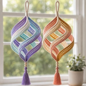 Puede incluir: Dos adornos colgantes en espiral hechos de tela de ganchillo. El adorno de la izquierda presenta un degradado de morado, blanco y azul claro. El de la derecha presenta un degradado de naranja, crema y amarillo. Cada uno tiene una borla y cuentas de madera.