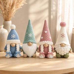 May include: Four crocheted gnome figurines with pointed hats. The first is blue with a snowflake, the second green with a snowflake, the third pink with white hearts, and the fourth cream with a pink snowflake and a textured white beard.