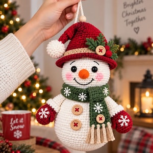 Può includere: Un ornamento a forma di pupazzo di neve all'uncinetto con un cappello rosso con pompon bianco, una sciarpa verde con ricami di fiocchi di neve e frange, muffole rosse con dettagli a fiocco di neve e due bottoni in legno sul corpo bianco.