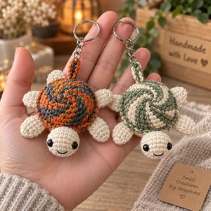 Può includere: Due portachiavi a forma di tartaruga all'uncinetto con anelli in metallo. Una tartaruga ha un guscio con motivo a spirale arancione e grigio scuro, l'altra ha un guscio con motivo a spirale verde salvia e crema. Entrambe hanno testa e arti color crema.