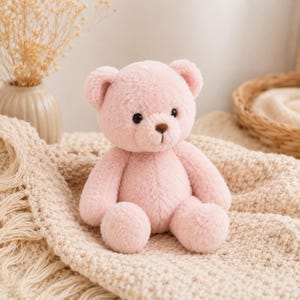 May include: A plush teddy bear with soft, light pink fur, round ears, and a small brown nose. The bear has two black, circular eyes and is seated in an upright position. Its limbs are rounded and its overall texture appears fuzzy and soft.