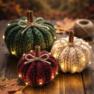 Peut inclure: Trois citrouilles crochetées de couleurs vert foncé, bordeaux profond et crème. Chaque citrouille possède une tige en bâton de cannelle, un nœud en ficelle et est entourée d'une fine guirlande lumineuse blanc chaud sur une surface en bois.