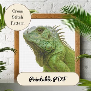 Puede incluir: Un patrón de punto de cruz enmarcado que presenta una iguana verde detallada. La obra de arte está en un marco de madera e incluye el texto "Cross Stitch Pattern" y "Printable PDF". La iguana está representada en tonos verdes.