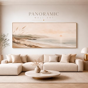 Könnte beinhalten: Ein panoramisches Wandbild mit einer Küstenlandschaft, Sanddünen, hohem Gras und einem ruhigen Ozean unter einem sanften Sonnenuntergangshimmel. Das Kunstwerk ist in einem goldenen Rahmen über einem beigen Ecksofa mit passenden Kissen platziert.
