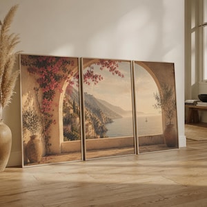 Könnte beinhalten: Ein dreiteiliges Wandbild-Set mit einer mediterranen Küstenszene. Die Drucke zeigen Steinbögen, leuchtend rosa Bougainvillea, Terrakottatöpfe und einen weiten Blick auf ein Küstendorf und das Meer in einem sanften, warmen Licht.
