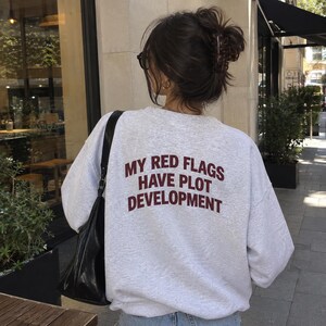 May include: A light gray crewneck sweatshirt featuring the dark red text "MY RED FLAGS HAVE PLOT DEVELOPMENT" printed on the back. The garment has long sleeves and a relaxed fit, worn with a black shoulder bag and blue denim jeans.