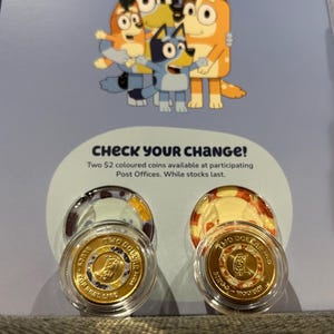 Puede incluir: Dos monedas doradas de $2 en estuches de plástico transparente, con una familia de dibujos animados. El texto "CHECK YOUR CHANGE!" está encima de las monedas. Las monedas son parte de una serie disponible en las oficinas de correos.