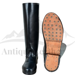 Puede incluir: Botas de montar de cuero negro, una de pie y la otra mostrando la suela. La suela es de cuero marrón con tachuelas metálicas. Las botas son de diseño clásico, adecuadas para equitación o eventos históricos.