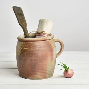 May include: A vintage ceramic jar with a lid, handle, and a wooden spoon inside. A rolled-up cloth tied with twine and a small red onion with green sprouts are also present. The jar has a pinkish-brown colour.