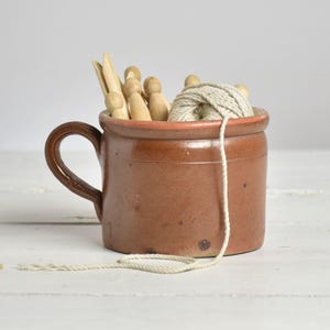 Może przedstawiać: Brązowy ceramiczny kubek wypełniony drewnianymi klamerkami do prania i kulą beżowego sznurka. Kubek ma uchwyt i lekko szorstką fakturę, sugerującą styl vintage lub rustykalny. Sznurek zwisa z kubka.