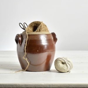 May include: A brown ceramic jar with two handles, containing twine and scissors. A ball of twine is next to the jar. The jar has a glossy finish and is set against a white backdrop.