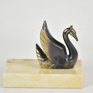 May include: A bronze swan figurine with gold-colored wings and a black body, perched on a rectangular, light-colored stone base. The swan has a curved neck and detailed feathers, creating an elegant decorative piece.