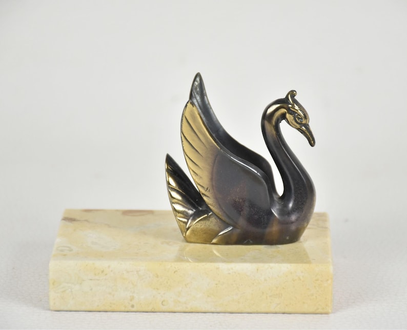 May include: A bronze swan figurine with gold-colored wings and a black body, perched on a rectangular, light-colored stone base. The swan has a curved neck and detailed feathers, creating an elegant decorative piece.