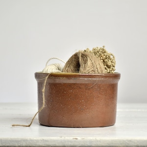 Op de afbeelding: Een bruine keramische pot gevuld met garen en gedroogde bloemen. De pot heeft een afgeronde vorm en een licht glanzende afwerking. Het garen is in tinten beige en bruin, en de bloemen zijn lichtbruin van kleur. Een stuk touw hangt uit de pot.