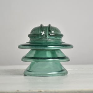 Peut inclure: Un isolateur électrique en verre vert sarcelle vintage présentant une conception à plusieurs niveaux avec un sommet arrondi et une base large et évasée. Le matériau translucide a une finition lisse et polie avec des crêtes moulées visibles.