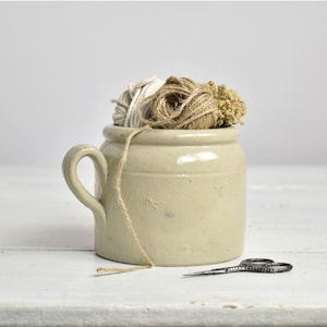 Puede incluir: Un tarro de cerámica beige con asa, lleno de ovillos de hilo y flores secas. Un trozo de hilo cuelga por un lado. Unas tijeras pequeñas y negras descansan sobre la superficie blanca frente al tarro. El tarro tiene una estética rústica y vintage.