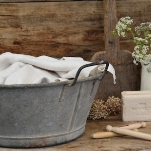 Może przedstawiać: Stara, ocynkowana metalowa wanna z białą szmatką w środku. Wanna stoi na drewnianej powierzchni, obok niej znajduje się mydło i drewniane spinacze do bielizny.