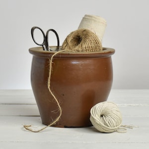 Peut inclure: Un pot en céramique marron rempli de ficelle, de tissu et de ciseaux. Une pelote de fil blanc est posée à côté du pot. Les objets sont disposés sur une surface en bois blanc, créant une esthétique rustique et vintage.