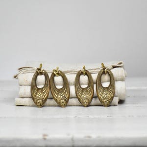 Op de afbeelding: Vier ovale ladehandgrepen van messing met een decoratief reliëf bloemmotief aan de basis en een gladde, afgeronde bovenlus. Elk stuk heeft een metallic gouden afwerking met subtiele donkere patina-accenten en een symmetrisch ontwerp.