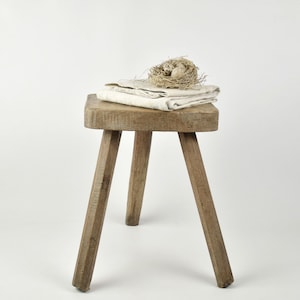 May include: A rustic, three-legged wooden stool with a square top. A folded, off-white cloth and a small bird's nest with eggs rest on the stool. The stool and nest are set against a plain white background.